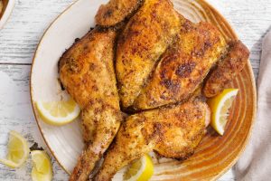 Spatchcock whole roasted chicken served on brown plate with lemon wedges.