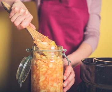 Woman stirring homemade sauerkraut in glass fermenting jar with wooden spoon.
