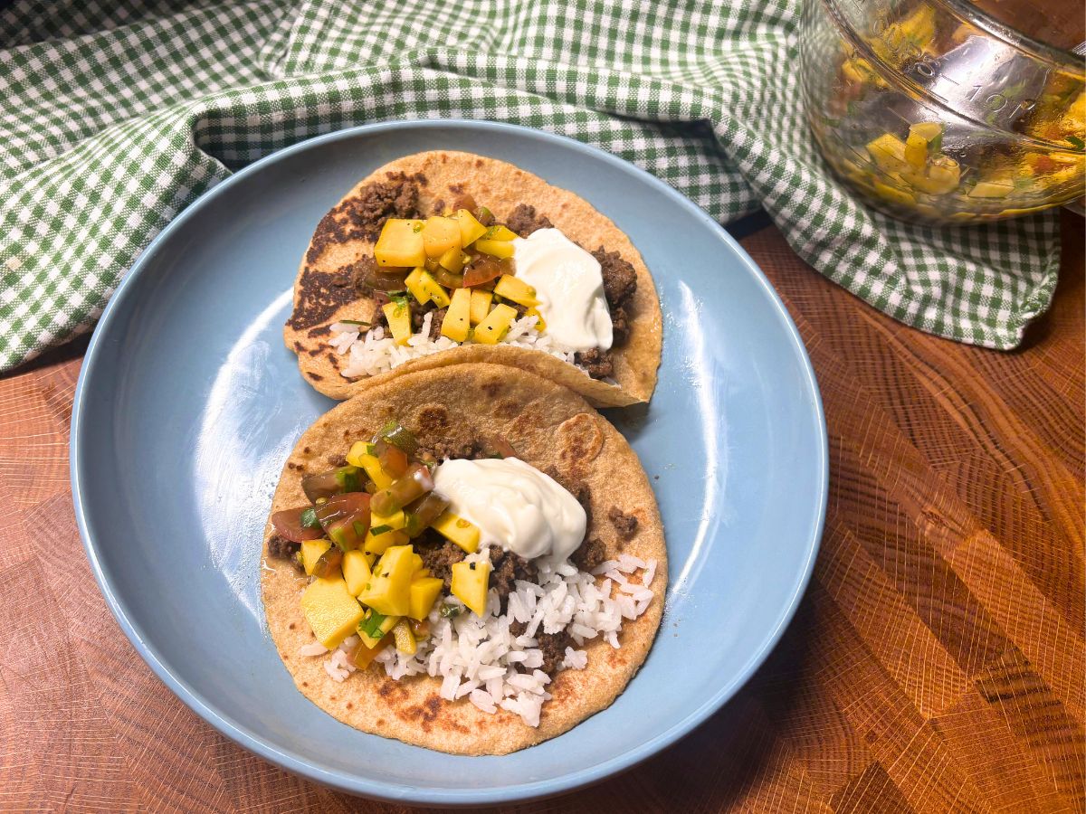 two beef tongue tacos with mango salsa cilantro lime rice, and sour cream on a homemade fresh milled flour tortilla on a blue plate sitting on a butcher block counter top