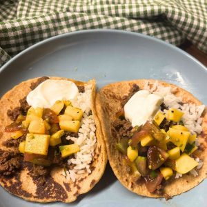 two homemade beef tongue tacos on a blue plate with mango salsa, rice, and sour cream sitting next to a green checked tea towel