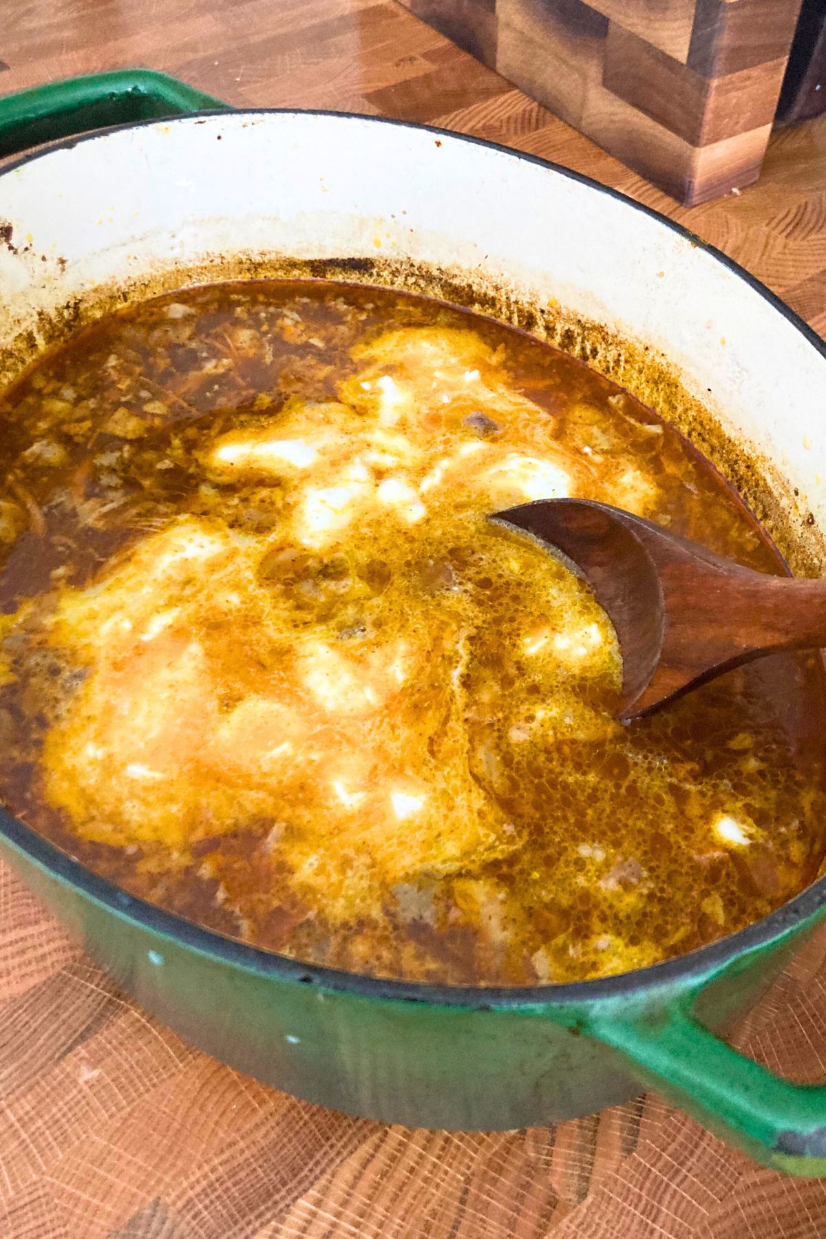 Lamp curry soup in a green dutch oven being stirred by a wooden spoon on a butcher block counter top