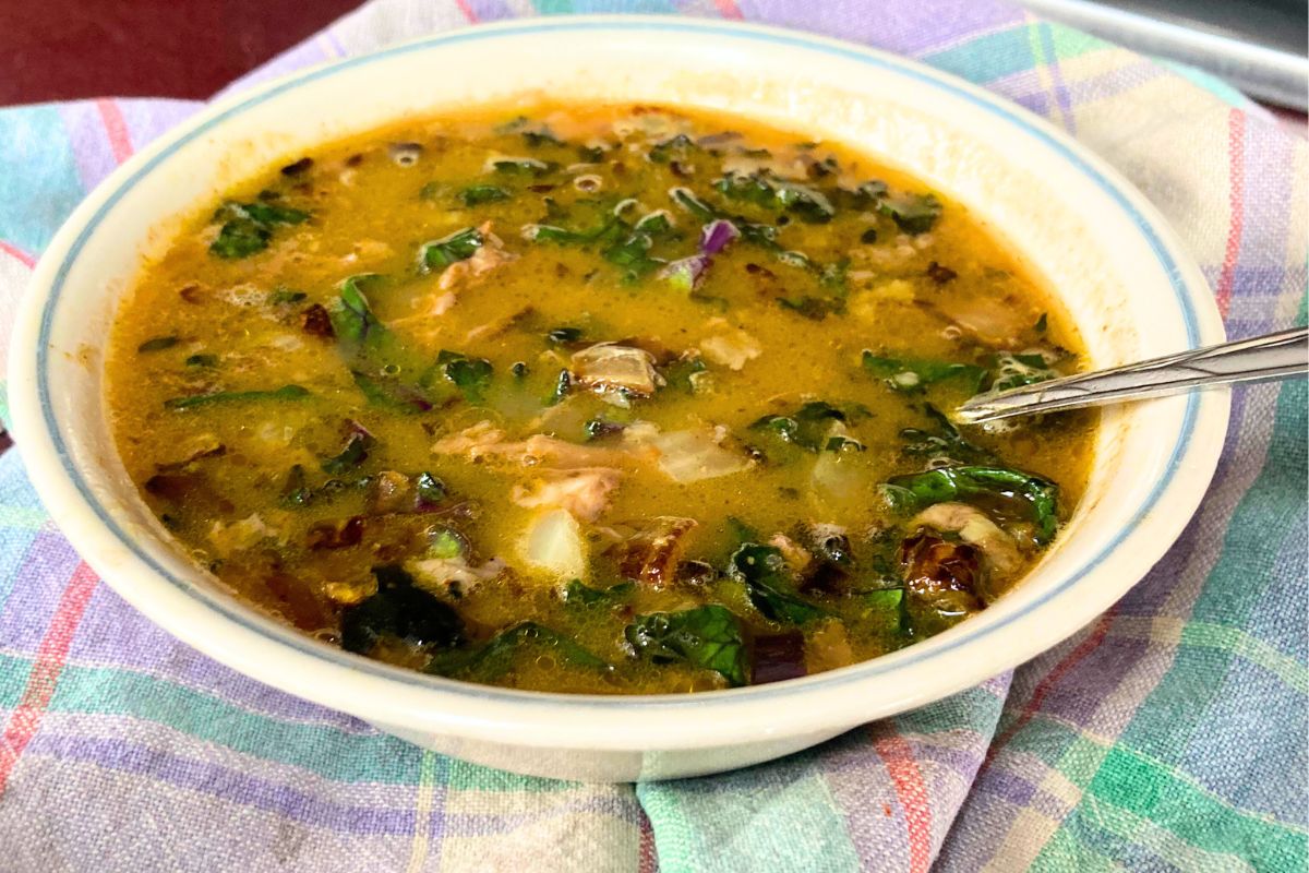 lamb curry soup in a white bowl with a blue rim and spoon on top of a purple and green plaid towel