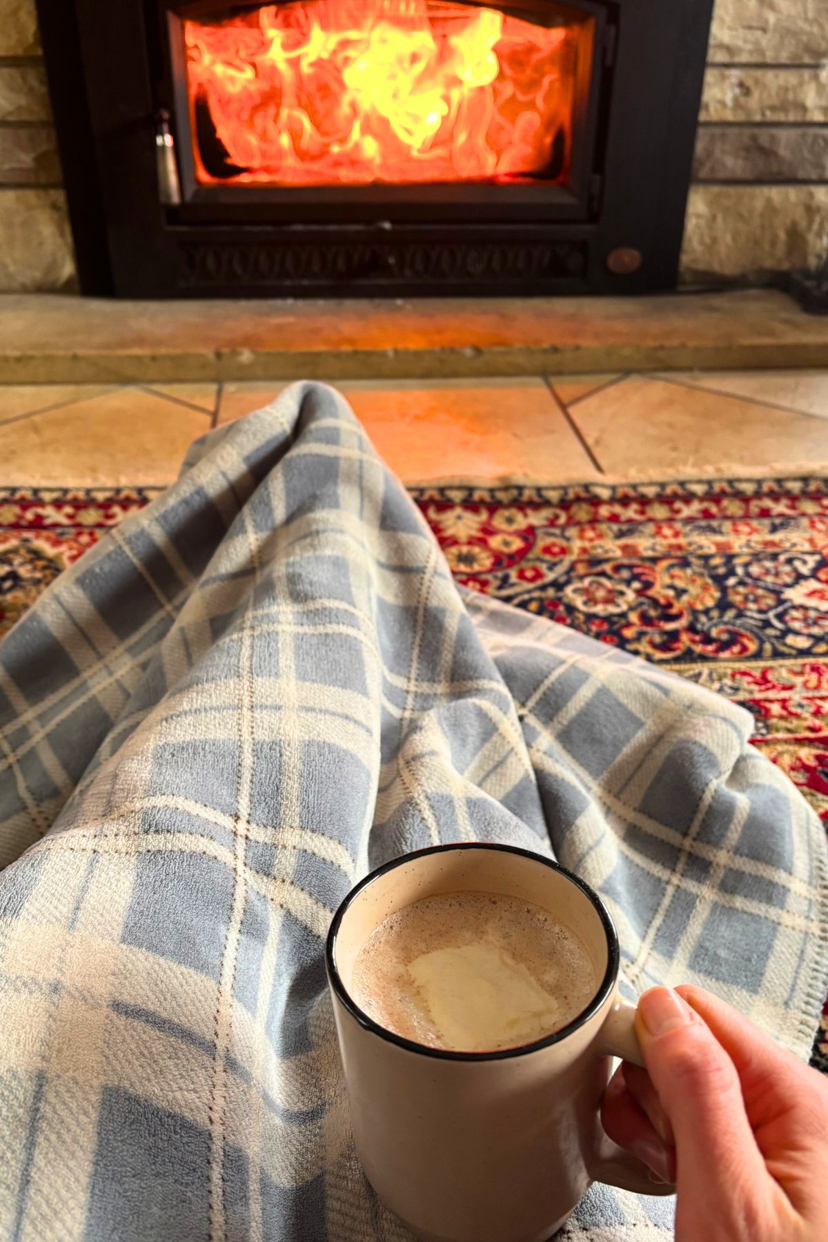 brown mug filled with a warm winter drink and topped with a homemade marshmallow being held by a hand on someone sitting on the floor with a blue and white plaid blanket in front of a wood burning fireplace