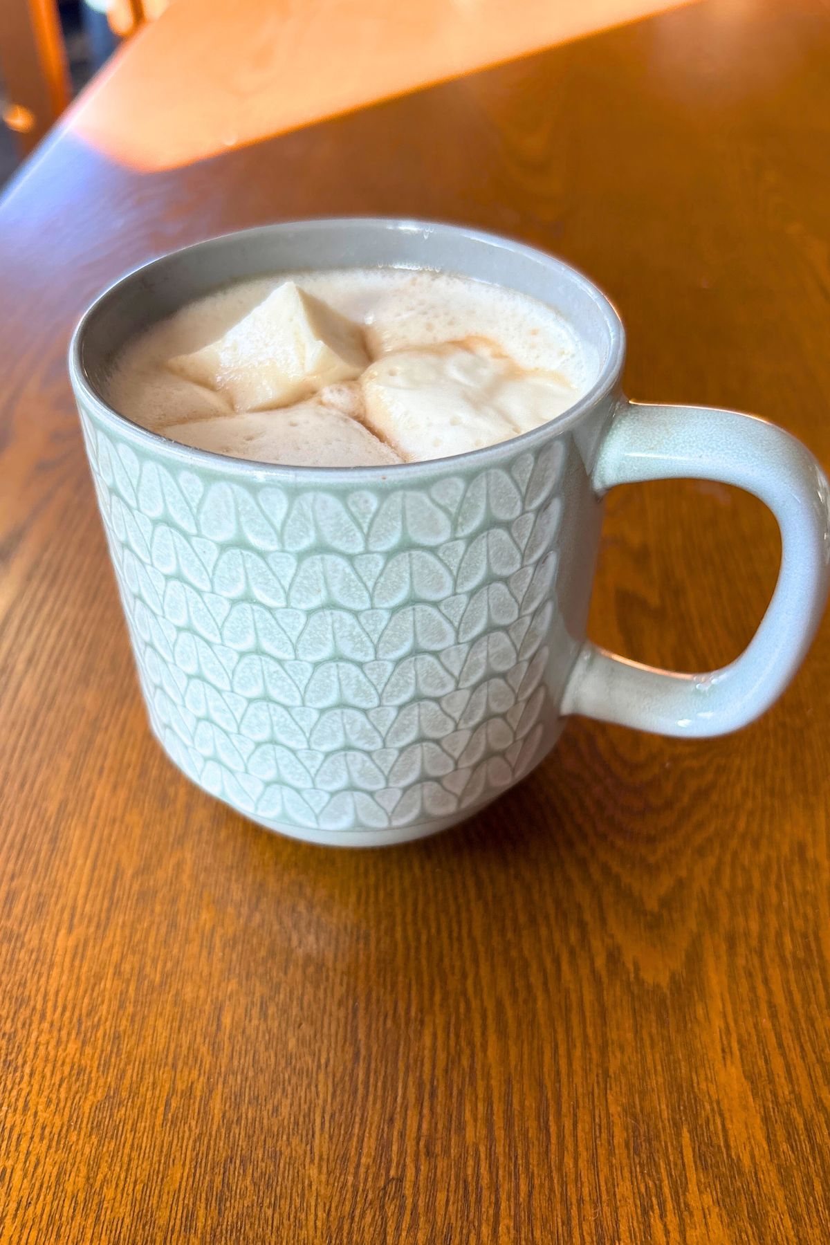 a warm winter drink which is hot cocoa with 3 homemade marshmallows on top in a grey and white patterned mug sitting on a wooden table