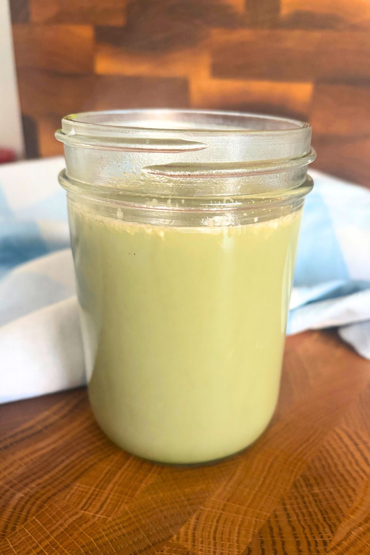 a warm winter drink which is a green matcha latte in a wide mouth pint-sized mason jar on a wooden counter top in front of a blue and white checked towel and a wooden cutting board