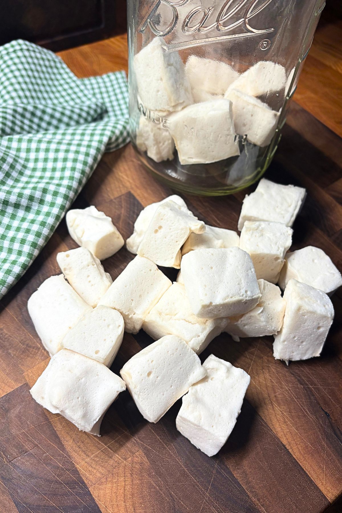 homemade marshmallow on a wooden cutting board in front of a mason jar of marshmallows next to a green and white checked towel