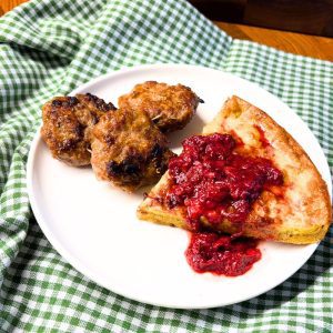 gluten free oven pancake with fresh milled flour on a white plate covered with strawberries with three pork sausage patties on top of a green and white checked towel