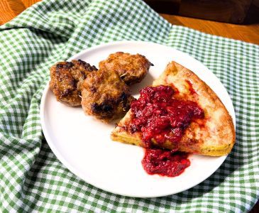 gluten free oven pancake with fresh milled flour on a white plate covered with strawberries with three pork sausage patties on top of a green and white checked towel