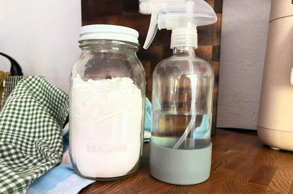 non-toxic cleaning products including mason jar filled with baking soda next to glass spray bottle of vinegar solution on a butcher block counter top