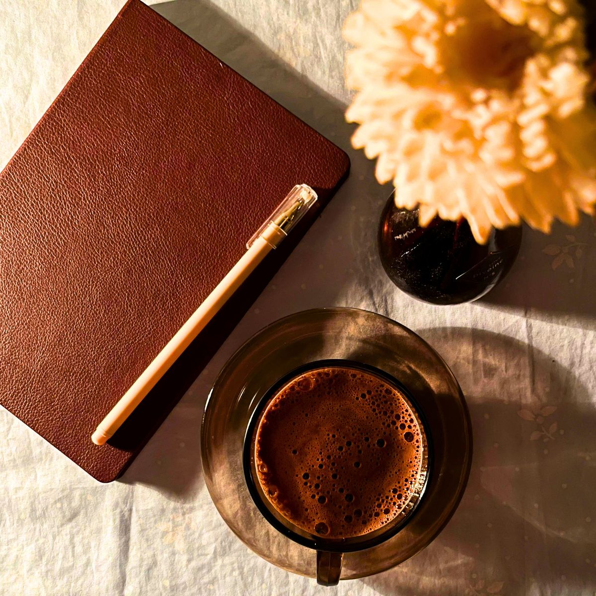 slow living brown leather journal next to a vase of yellow flowers and a mug of hot chocolate sitting on a white table cloth