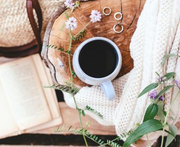 cup of coffee on a raw wood table next to a journal bag and white knit blanket