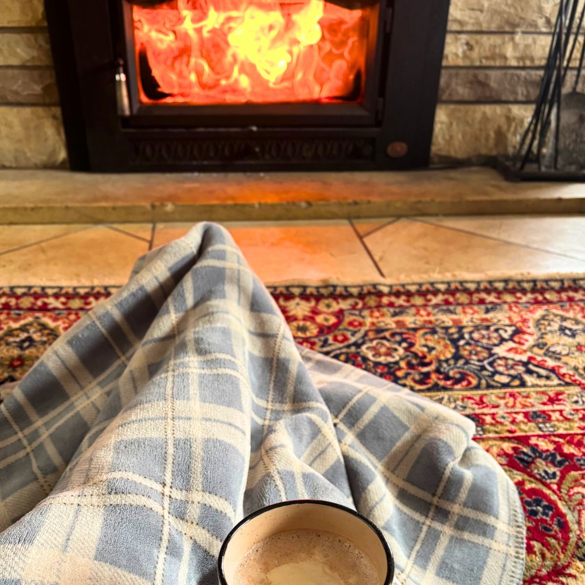 woman under a blanket with a hot chocolate with marshmallow sitting on the floor in front of a fireplace