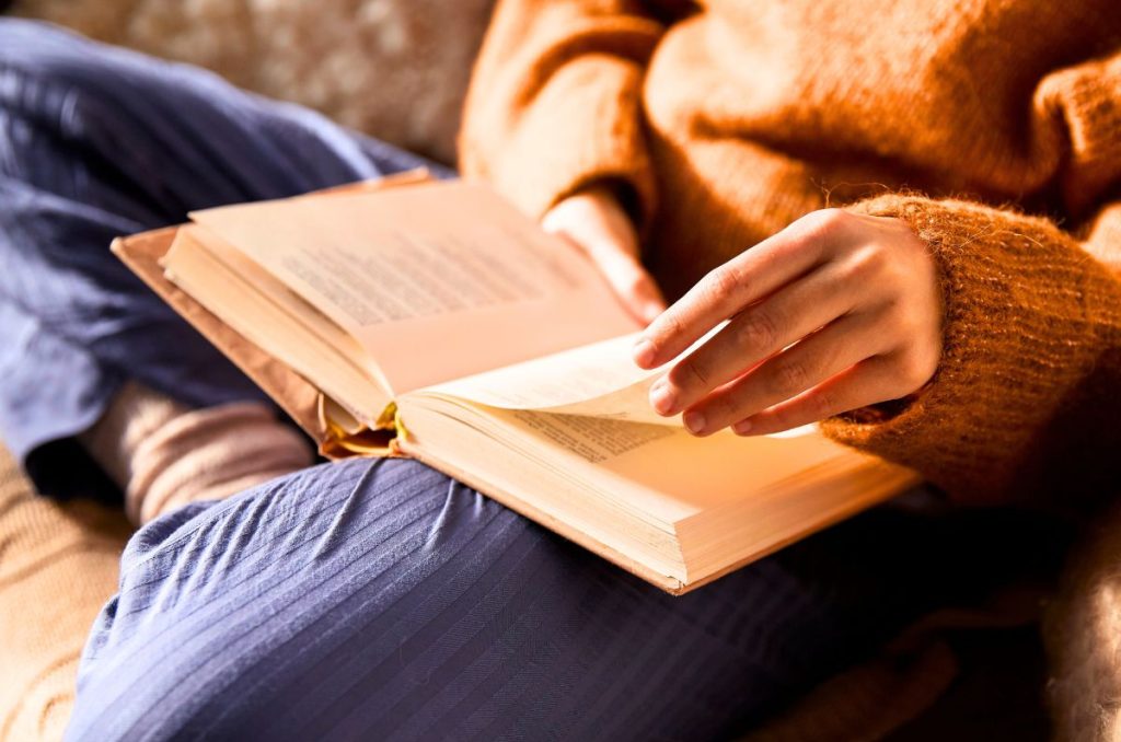 woman reading a book cross-legged on the couch wearing blue pants and a terracotta colored sweater