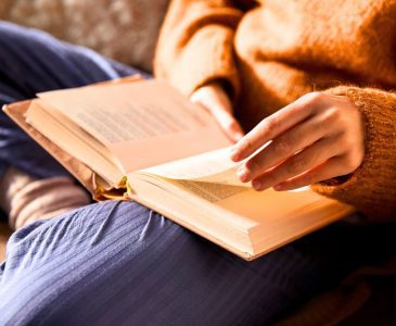 woman wearing a teracotta orange sweater and blue pants sitting on couch in a minimalist home reading a book