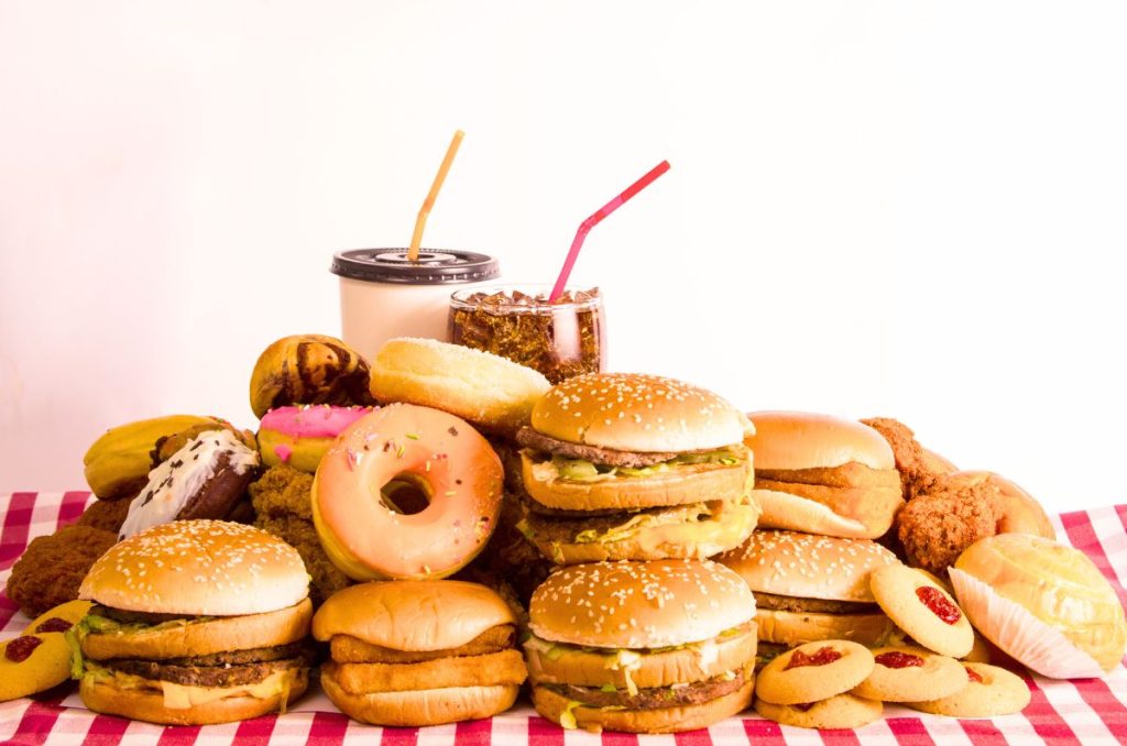 variety of fast foods including burgers donuts cookies sodas and fried foods all piled up on a red checked table cloth