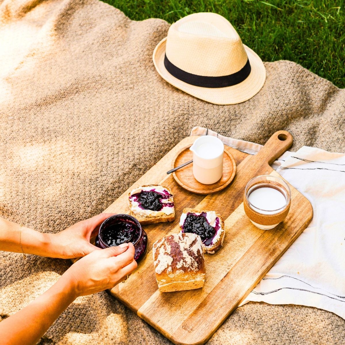 slow food picnic brunch of homemade bread, jam, and cream cheese with classes of milk on a wooden cutting board on a brown blanket in the grass
