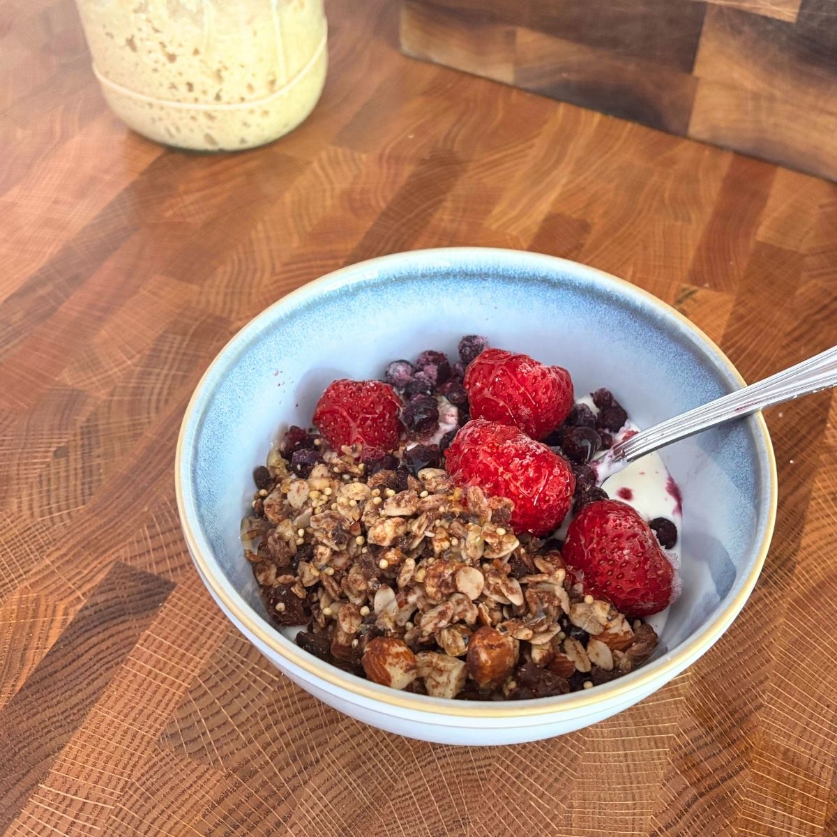 healthy bowl of yogurt, berries, homemade granola with a spoon in it on a butcher block counter top with sourdough starter in background