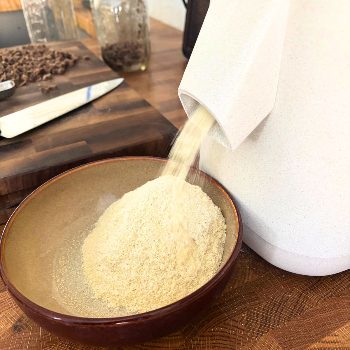 fresh milled flour coming out of a white grain mill into a brown bowl on a butcher block countertop