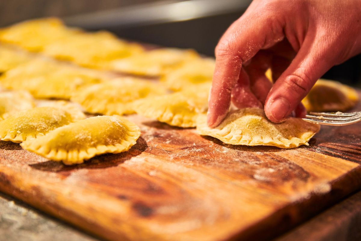 homemade raviolis on a wooden cutting board with a hand shaping a ravioli