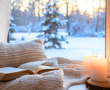 book on a cozy white chair with a white fleece blanket and two lit candles next to a wintery window
