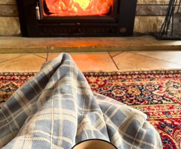 woman under a blue checked blanket with hot chocolate in hand in front of a roaring fire in her minimalist home