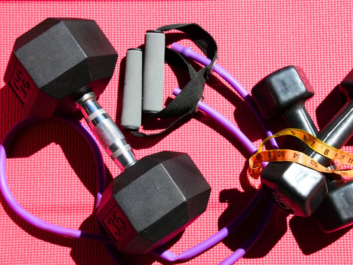 three dumbbells a purple exercise band with handles and a measuring tape laying on a pink exercise mat