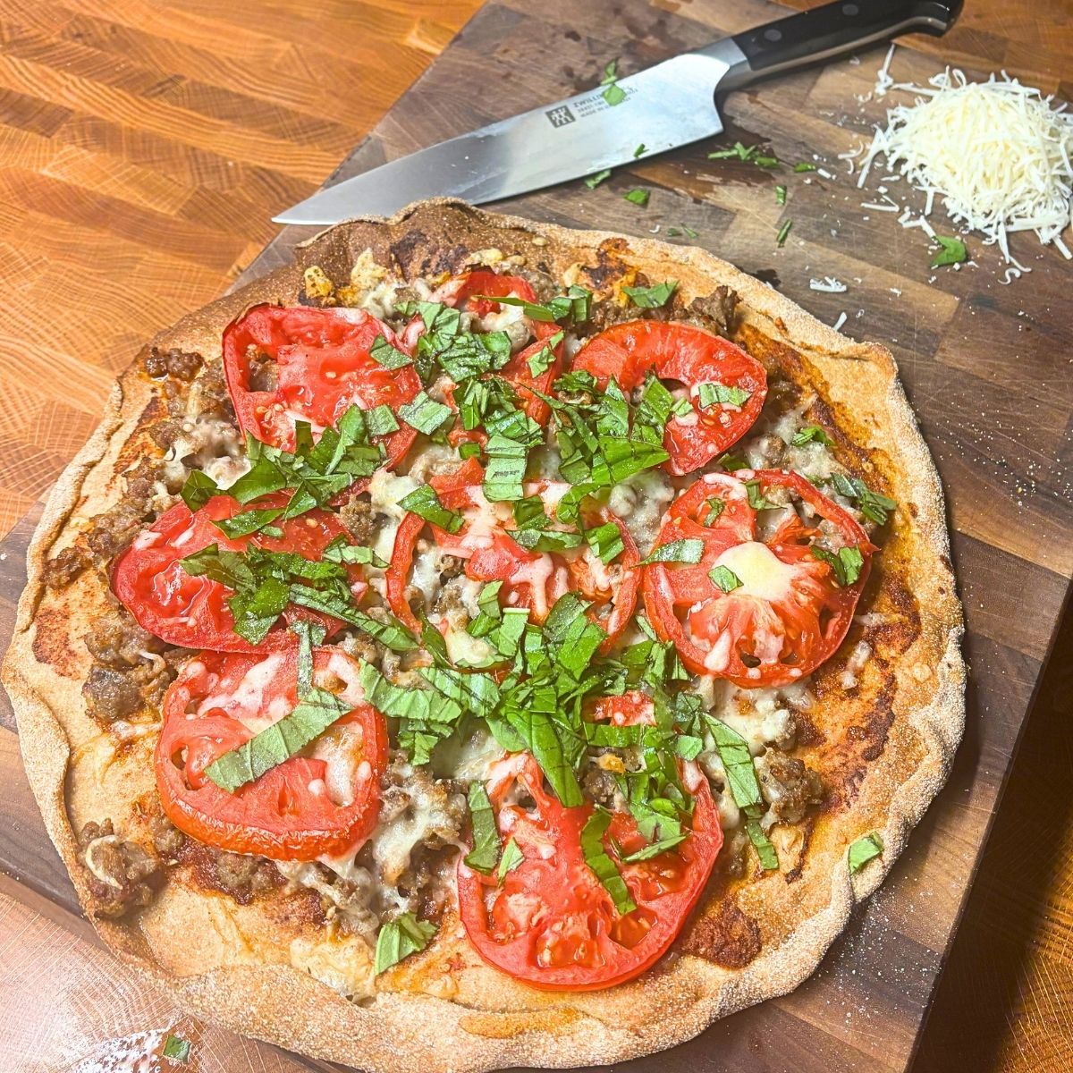 homemade pizza with sausage and tomato slices on a wooden cutting board next to a chef's knife and shredded cheese