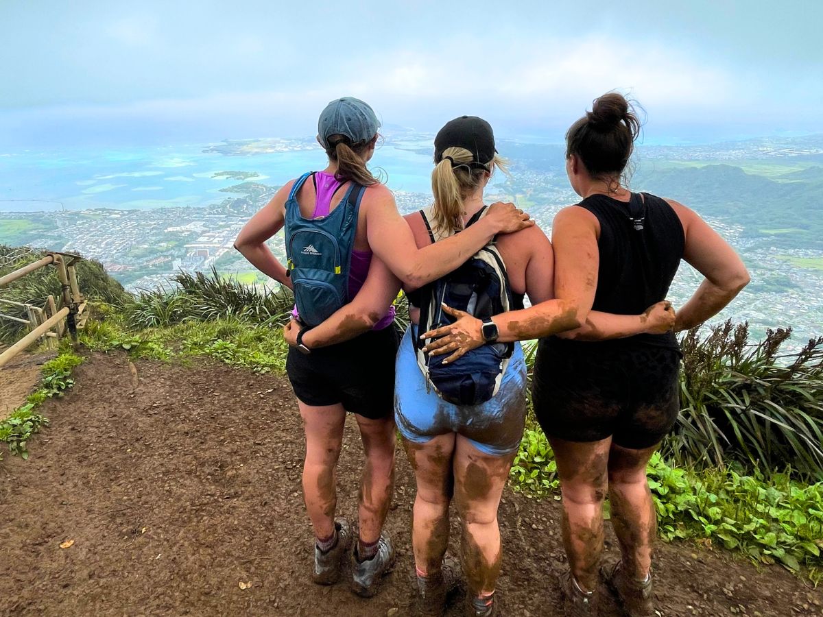 three young women standing facing away from the camera with their arms locked behind each others back looking over a Hawaii landscape from the top of a mountain