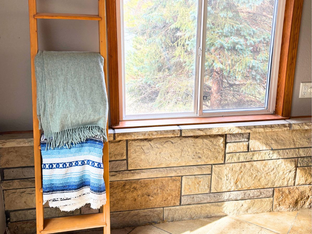 wooden ladder with blankets hanging on it next to a window above a stone wall