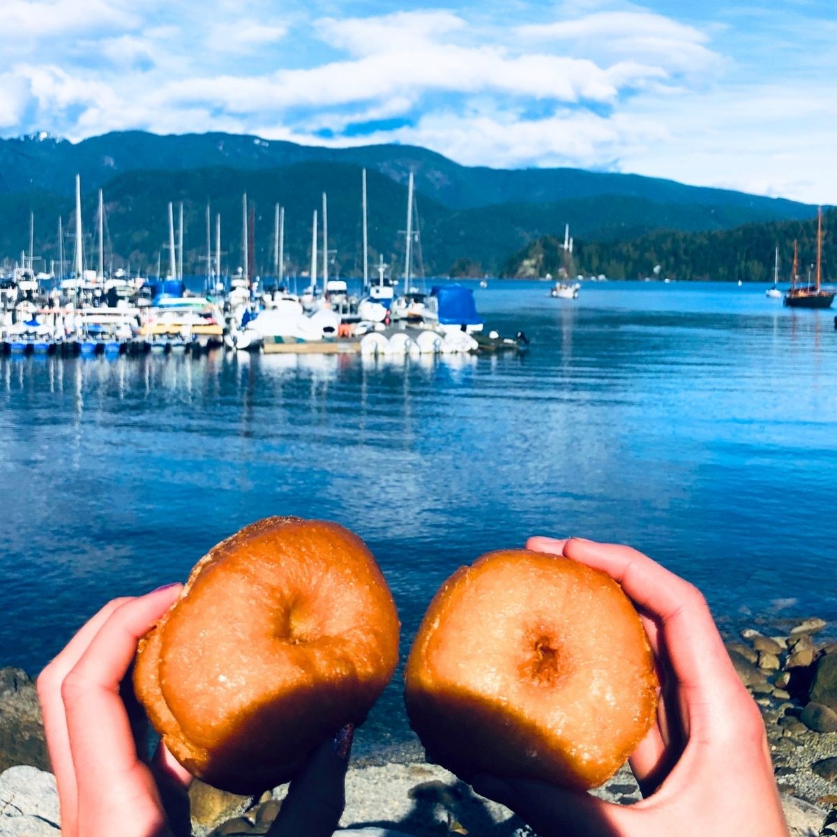 hands of two woman holding freshly made donuts in front of a blue harbor with boats