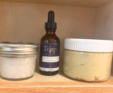 two glass jars with an opaque glass bottle between them on a wooden shelf containing non-toxic skincare products