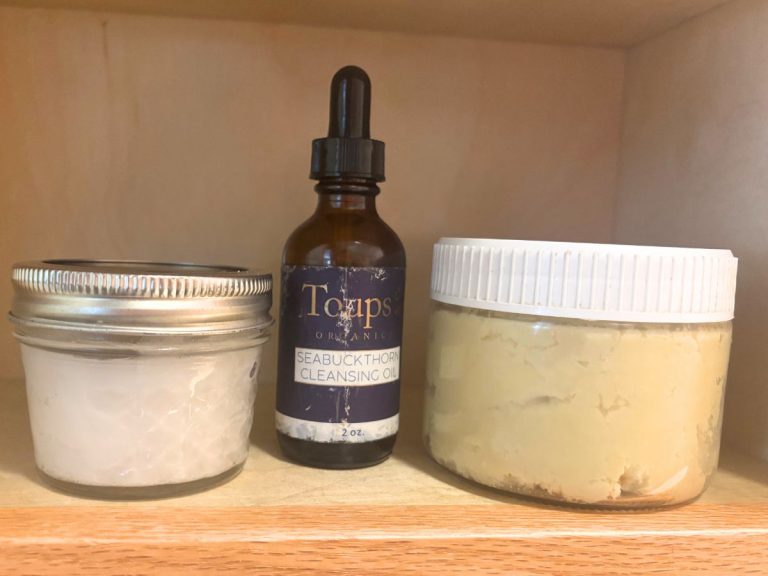 two glass jars with an opaque glass bottle between them on a wooden shelf containing non-toxic skincare products
